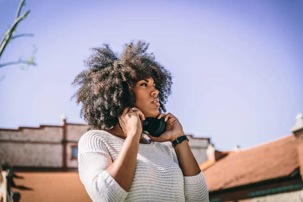 A brunette walks through a park while holding headphones wearing a white sweater.