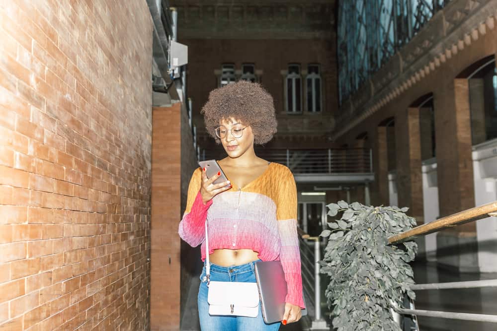 Una mujer afroamericana feliz con cabello oscuro y ropa informal revisa su teléfono dentro de una estación de tren.