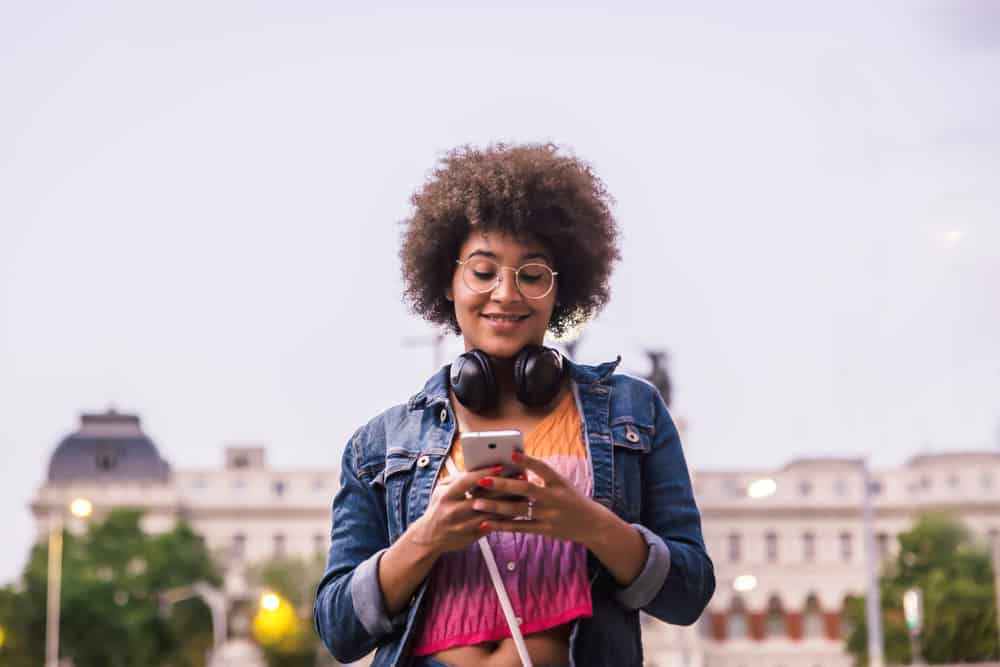 uma mulher com pele escura usando fones de ouvido pretos sorri para seu smartphone na rua vestindo uma camisa com cores vibrantes.