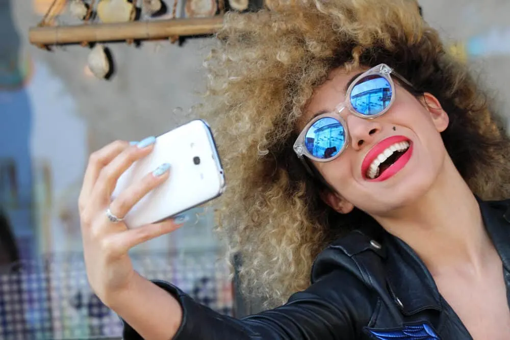 Mixed female smiling while taking a selfie wearing curly hair strands with brassy undertones.