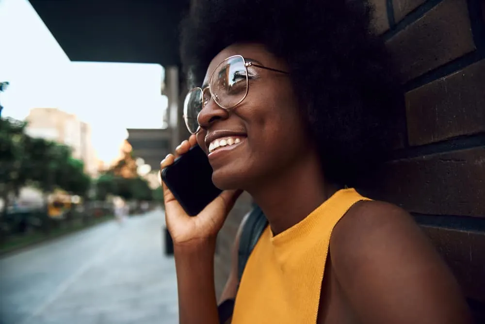 Cute black woman with naturally curly hair in glasses talking on the phone while leaning against a brick wall.