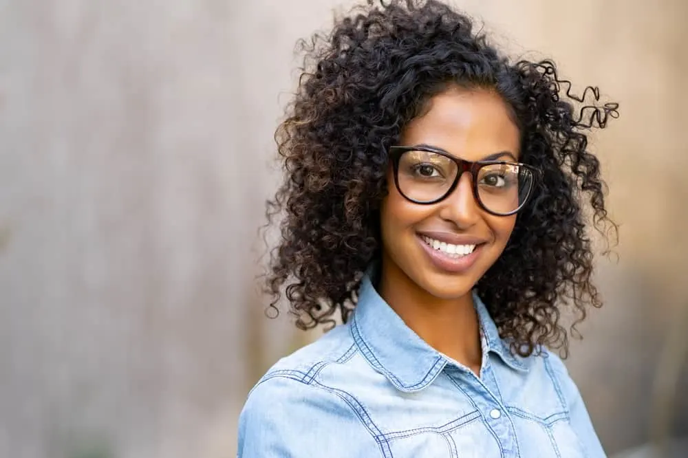 A cute young African American female is smiling while wearing black eyeglasses, a denim shirt, and neutral lipstick.