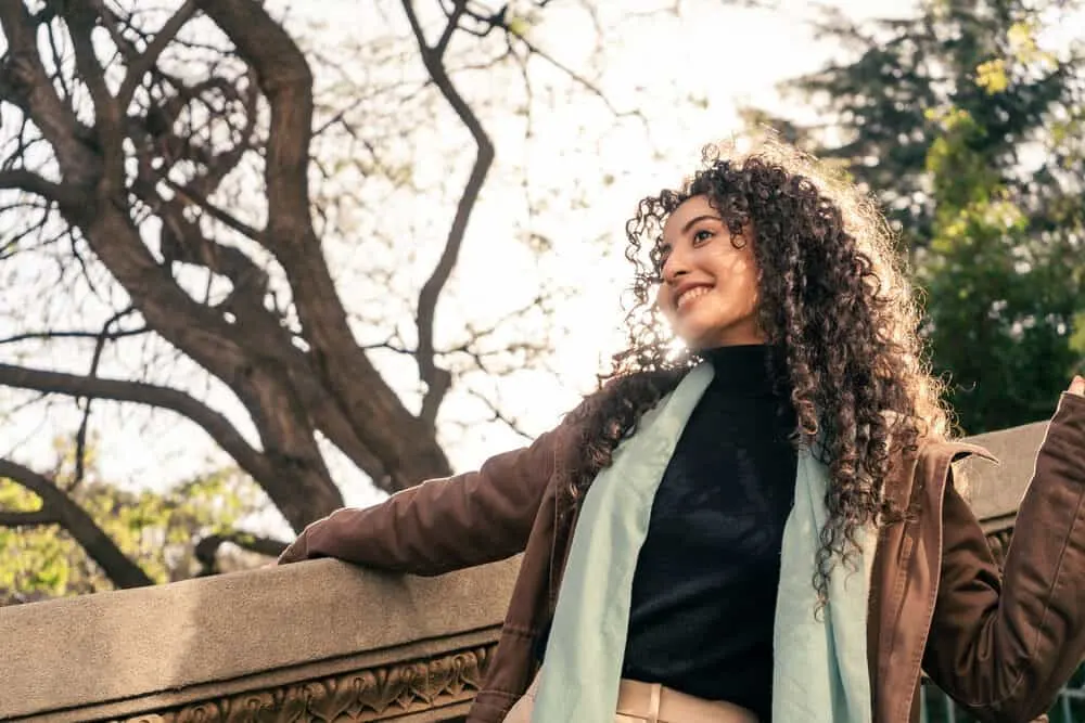 A lovely young and cute Hispanic woman with wavy hair leans against the concrete railing of an ancient staircase with moisturizing lotion on her face