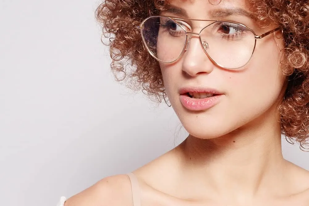 Young Caucasian woman wearing gold-framed glasses with darker hair and light skin-toned markup