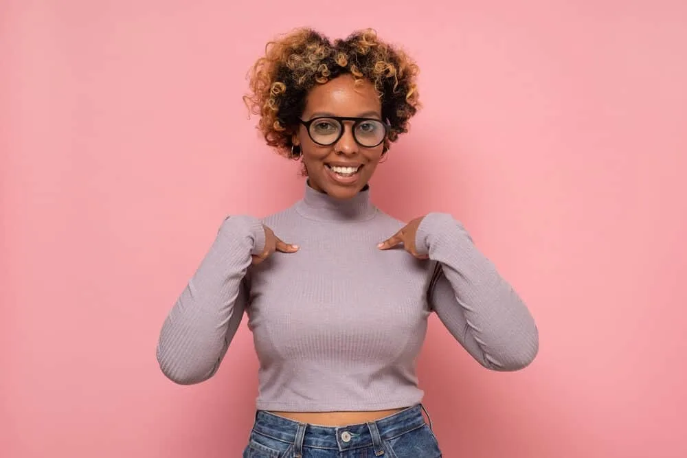 Black girl pointing fingers towards herself wearing a gray shirt with yellow brassy tones within her hair