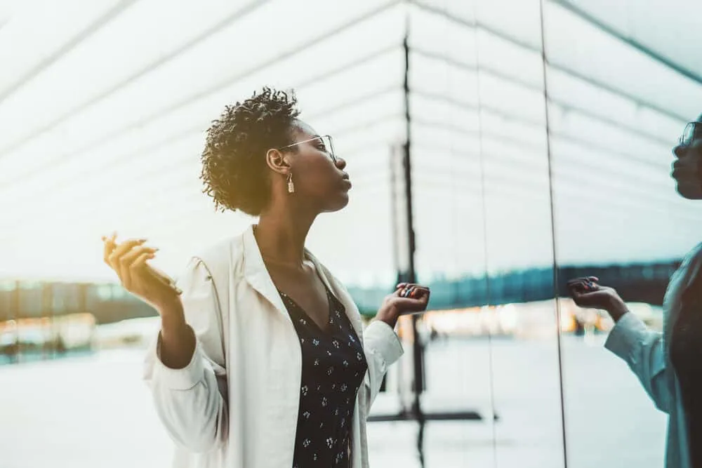 Black female with natural hair starting locs standing outside looking at her reflection in a glass building