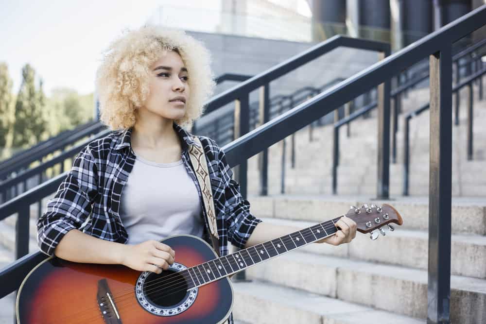 Schöne junge schwarze Dame spielt Gitarre mit aschblondem Haarfärbemittel, das ihr natürliches dunkles Haar bedeckt