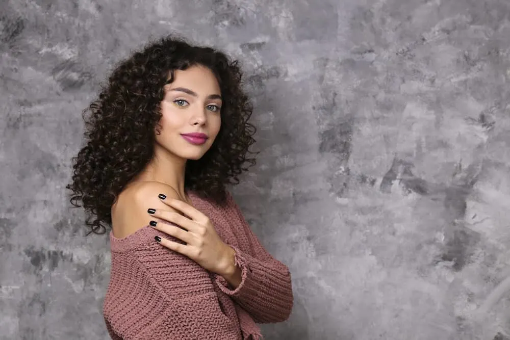 White girl with curly brown hair with brassy tones wearing a light purple sweater