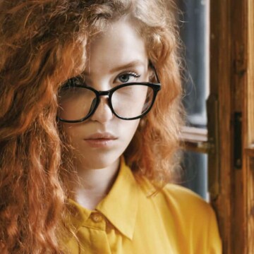 Girl with ginger highlighted hair standing near a window wearing a yellow dress shirt and black eyeglasses.