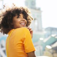 Happy black girl that's a hair care enthusiast with curly hair follicles wearing an orange shirt.