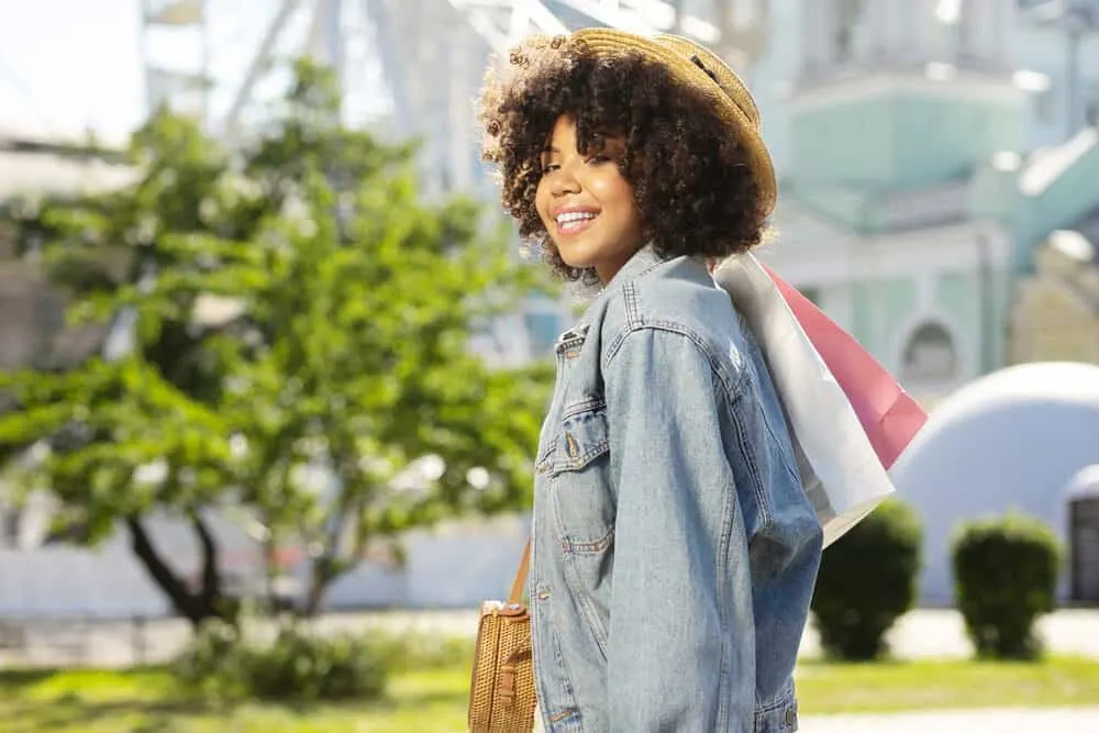 Adorable curly lady with a straw hat being delighted with shopping and smiling into the camera while carrying bags.