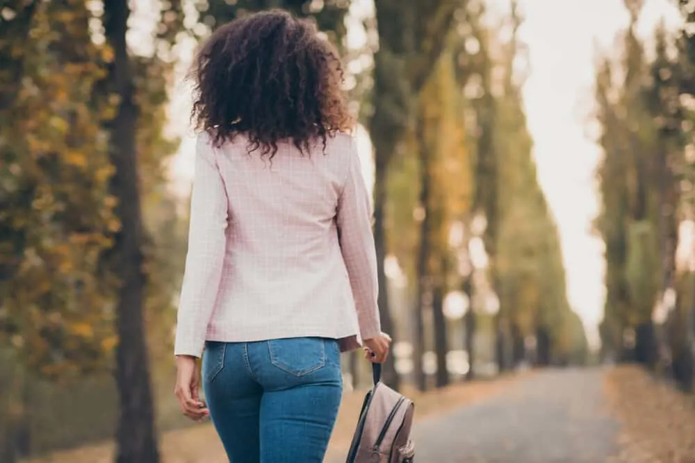 Adult female walking down a long road wearing a pink business jacket and blue jeans.