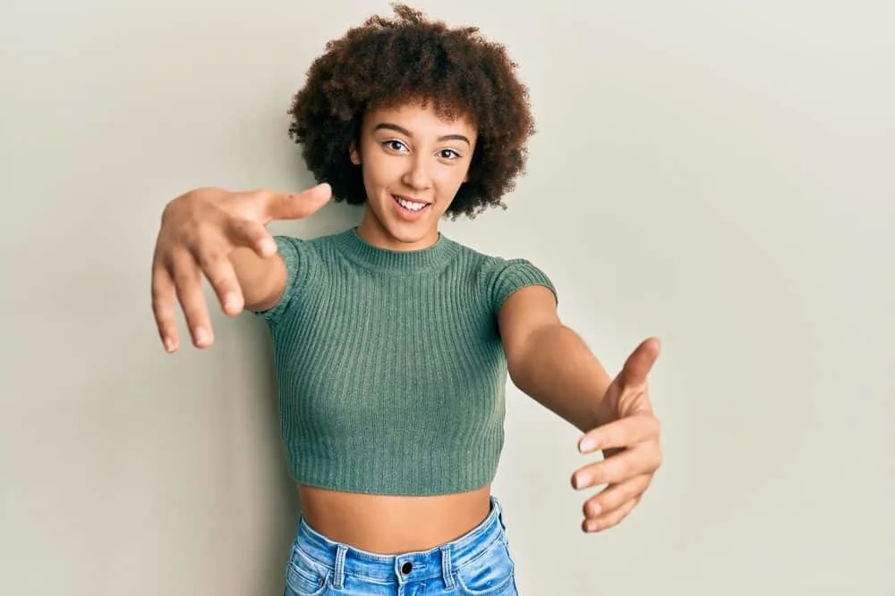 Young female with super curly long hair wearing a green sweater and blue jeans.