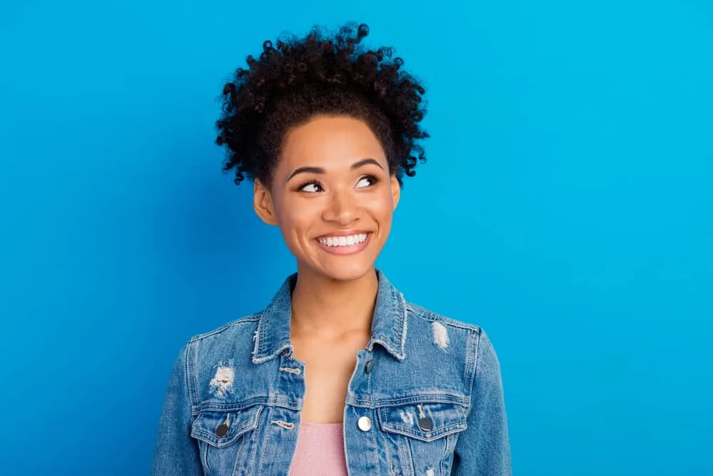 Black lady with dark brown curly dyed hair with a big smile wearing a blue jean jacket and pink shirt.