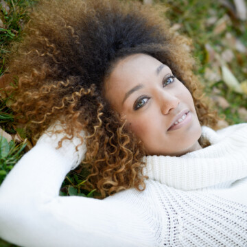 An African American woman with naturally dark hair suffering from hot roots and relaxing on the ground outside.