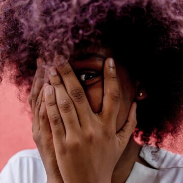 African American female with freshly washed hair covers face with hands.