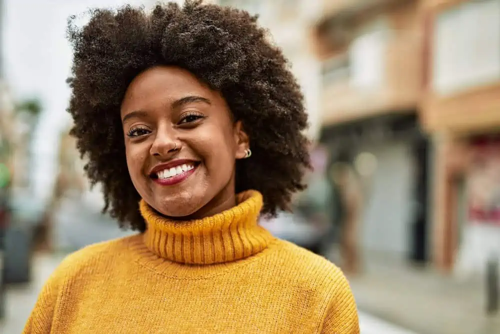 An African American lady with natural dark strands and gray roots wearing a yellowish-orange sweater.