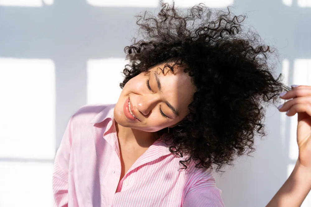 A lady with flawless skin and thick hair running her fingers through her curls and admiring her hair's health. 