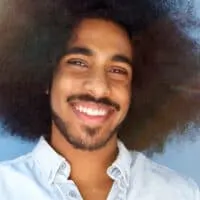 A smiling young man with facial hair and a full head with shoulder-length curls in their natural texture.