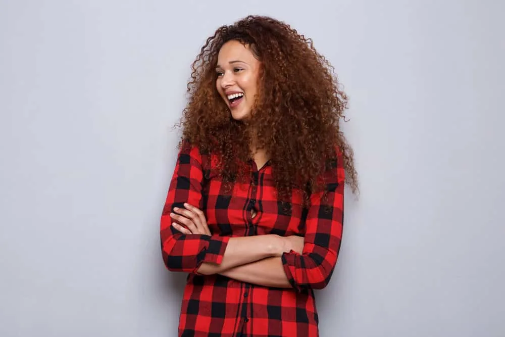 A young woman with natural curls dyed with demi-permanent hair color has her arms folded while laughing.