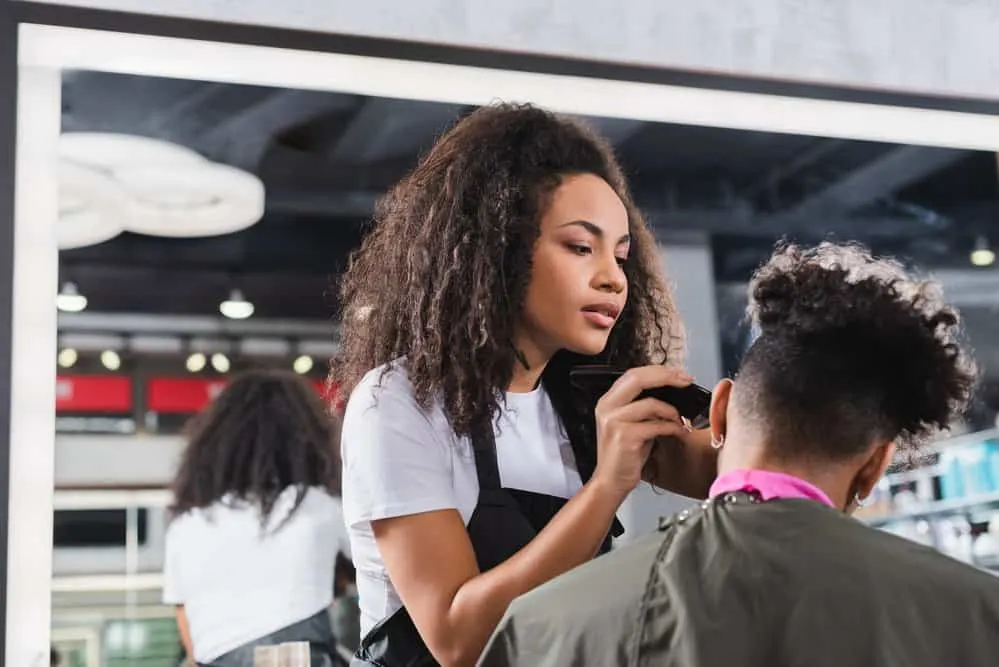 A lady with long hair that was a work-at-home stylist is cutting a neighbor's hair at a cosmetology school.