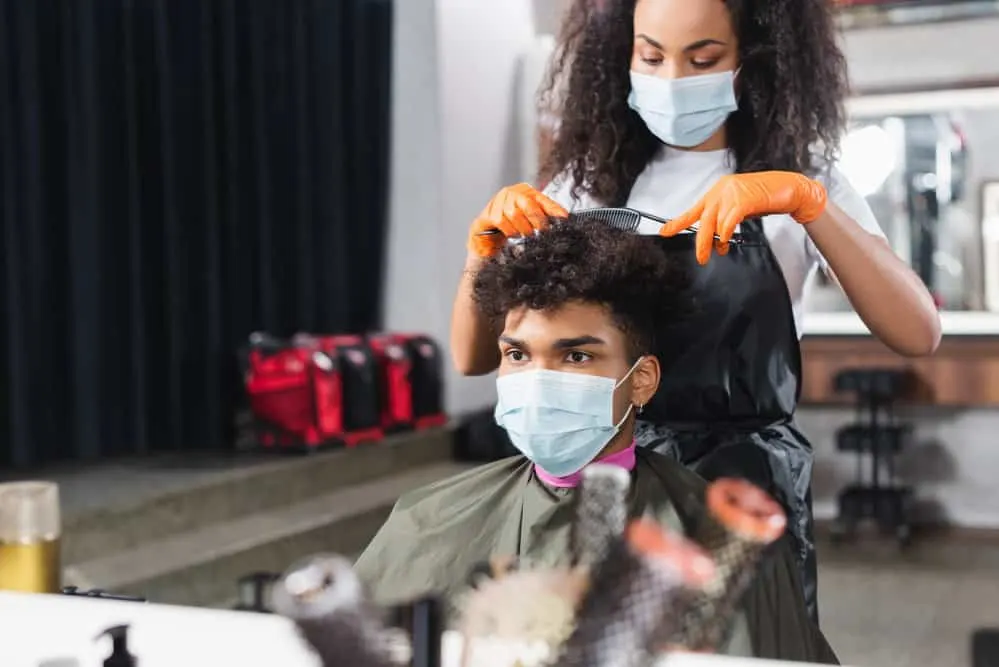 African male following a blow dry session is getting a hair cut from a female hairstylist with a comb and scissors.