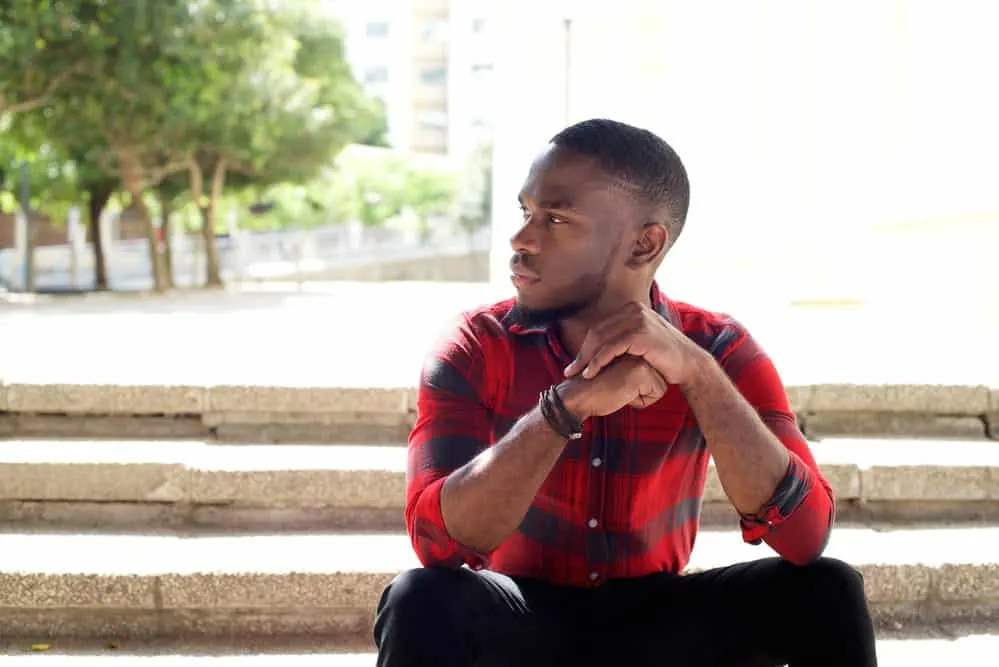 A calm young black male wearing a very short hair cut on his type 4 hair type is sitting outside relaxing.