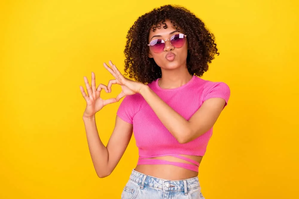 A female making a heart symbol with her hands is wearing fresh curls washed with shampoo and conditioner.