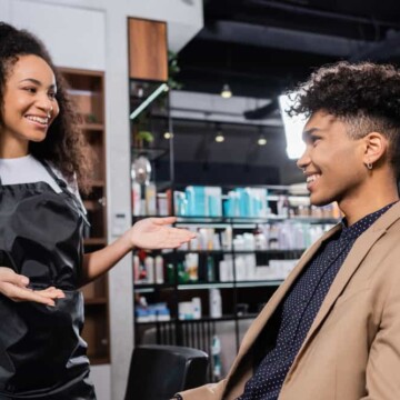 A man is sitting in a salon chair discussing his next hair appointment and about to give his professional hairstylist a tip.