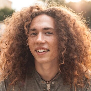 A young white male with curly, fine hair is wearing a shaggy wolf cut styled with texturizing spray on thicker hair.