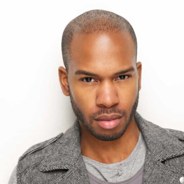 An African American man with thin hair and a receding hairline wearing a taper fade and a thick beard.