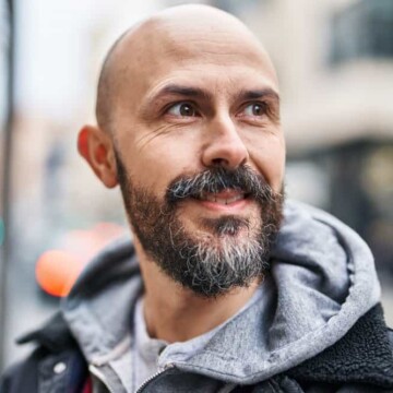 A young man with thinning hair follicles is sporting a bald head with grey hair in his thick facial hair.