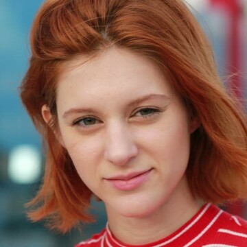 A super stylish young woman with red straight hair wearing a short hairstyle with blunt bangs styled with an organic holding spray on her oval face shape.