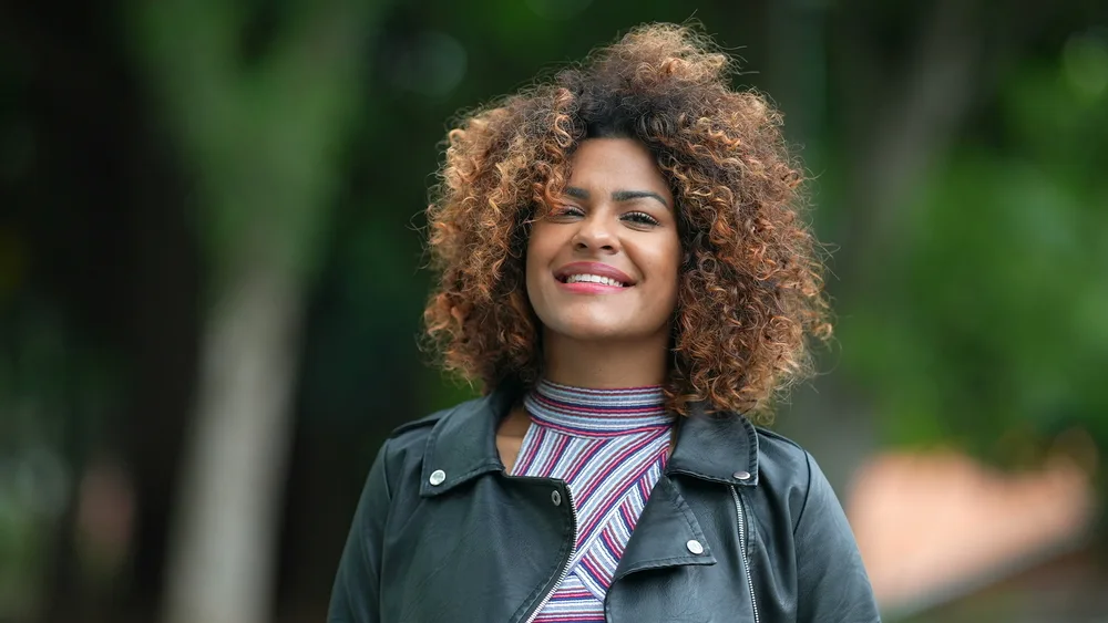 A captivating black female with warm skin tones showcases her colored hair with warm shades of strawberry blonde and golden brown for a natural look.