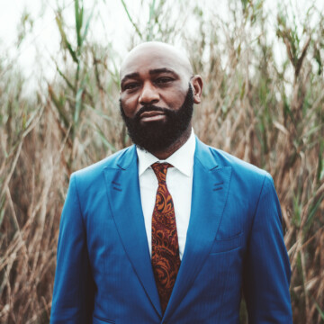 A handsome African man with a shaved head due to balding hair on his head and thick, dark brown facial hair creating a very masculine hairstyle.