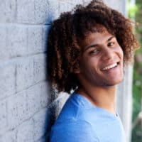 A young black man with fine hair is wearing his curly long hair in a classic style that resembles a dark brown and tan ombre hairdo with natural curls.