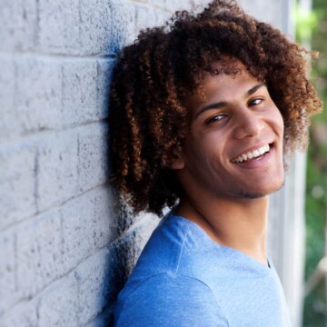 A young black man with fine hair is wearing his curly long hair in a classic style that resembles a dark brown and tan ombre hairdo with natural curls.