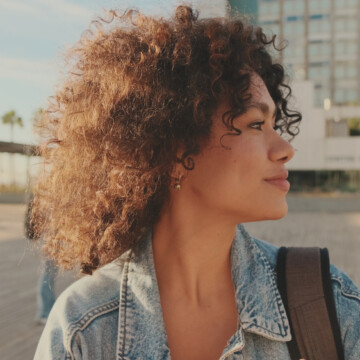 A black woman with a cute, confident smile proudly shows off her natural curls styled with shea butter and natural oils, like coconut oil, to hydrate her frizzy hair strands.